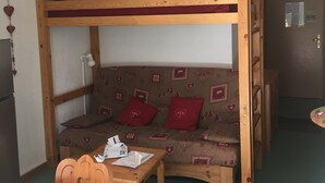 Een stapelbed met een houten frame en een slaapbank eronder. Er staat een klein tafeltje met een stoel voor de slaapbank.
