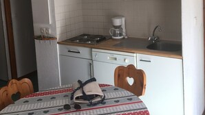 Een kleine keuken met witte kastjes, een magnetron, een koffiezetapparaat en een spoelbak. Er staat een tafel met een stoel in de vorm van een hart en een klein tafelkleed.