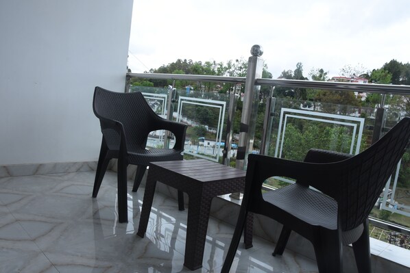 Varanda com duas cadeiras e uma mesa, guarda-corpo de vidro e vista para edifícios e árvores.
