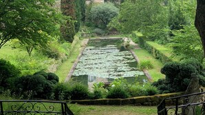 Un bâtiment en pierre, surmonté d’un toit de tuiles, entouré d’une végétation luxuriante et bordé d’un étang où flottent des nénuphars.