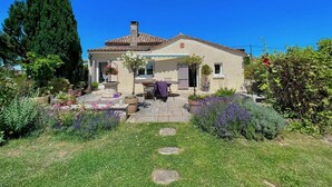 Une maison à deux étages, avec un toit en tuiles, un porche à l’avant et un jardin où poussent différentes plantes et fleurs.