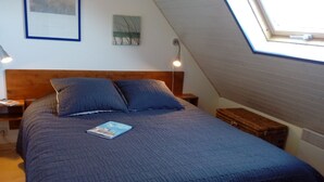 Camera da letto con soffitto a sbalzo, una testiera in legno, una coperta trapuntata blu, due cuscini, un comodino con una lampada, un piccolo comodino con un libro e una finestra con vista esterna.
