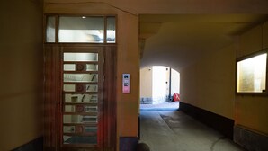 Un corridoio stretto con una porta in legno dotata di pannelli in vetro decorativi. Un piccolo pilastro in pietra si trova davanti alla porta. Il corridoio conduce verso una zona luminosa con un oggetto rosso in lontananza.