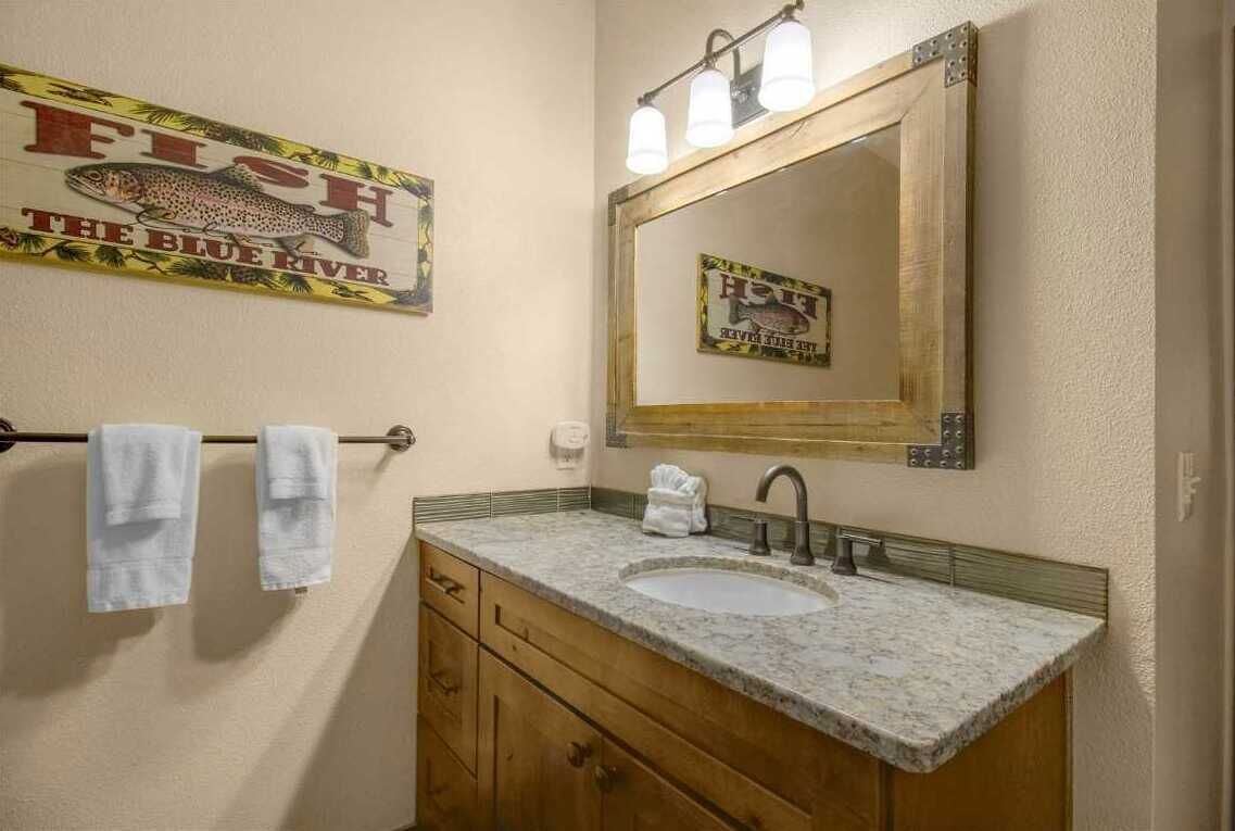 Baño con tocador de madera, lavamanos y espejo. Hay un perchero con dos toallas y un cartel sobre el espejo que dice 'FISH THE BLUE RIVER'.