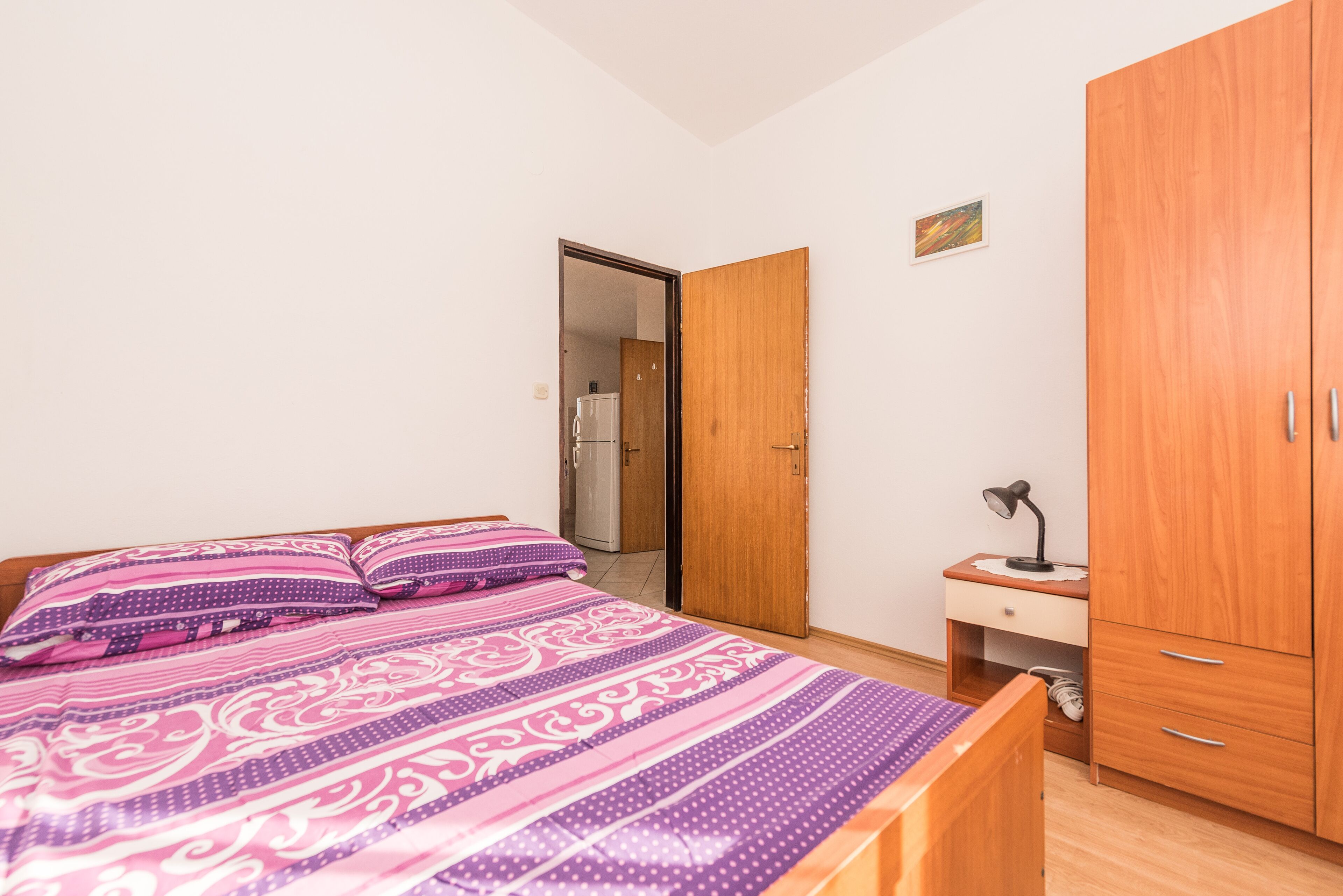 A bedroom with a bed, wooden wardrobe, and a nightstand with a lamp.