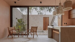 A modern interior with a dining area, a kitchen counter, and a large window overlooking a stone wall and trees. - Entorno Villas  (Tulum)