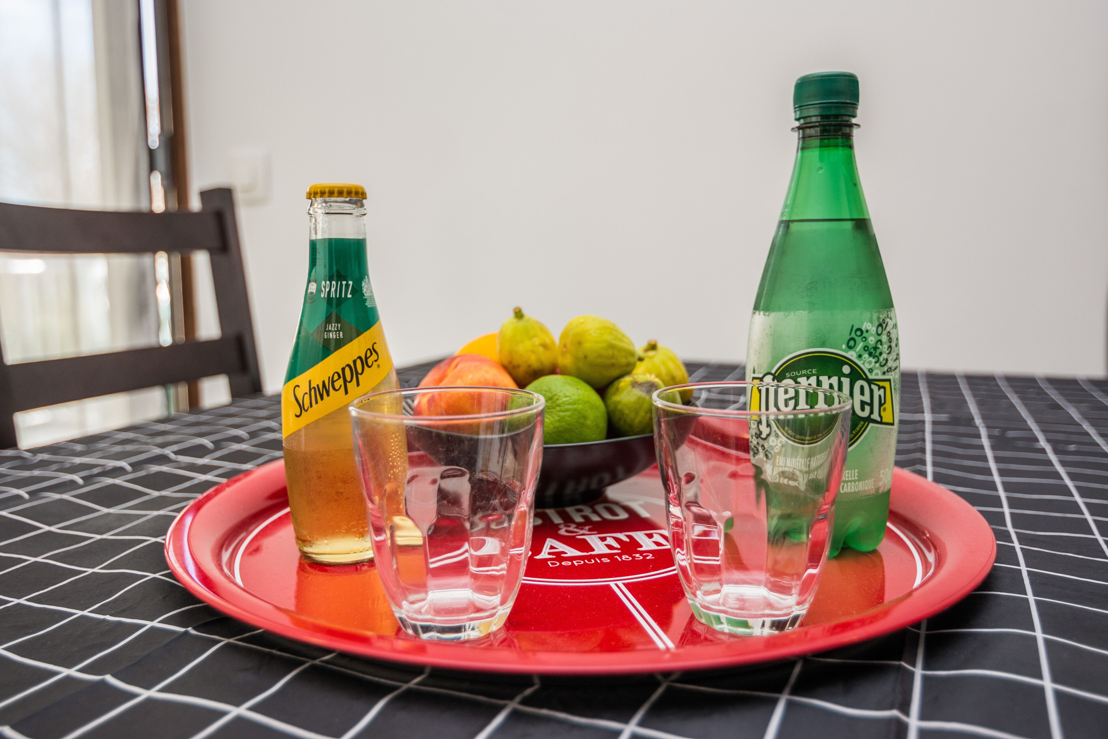 Una bandeja con una botella de Schweppes, dos vasos para chupitos y una botella de Menter sobre una mesa con una bandeja roja.