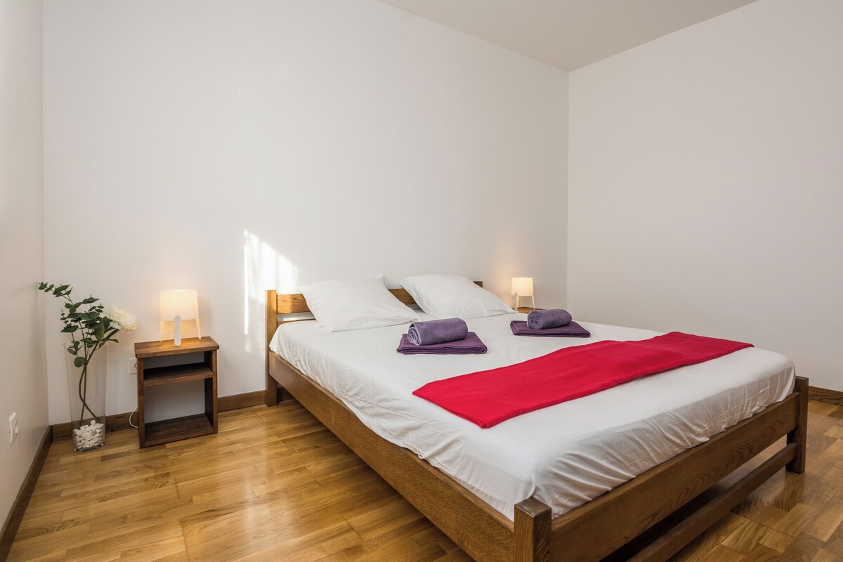 A neatly made bed with white linens and a red blanket, flanked by two bedside tables with lamps, in a room with wooden flooring.