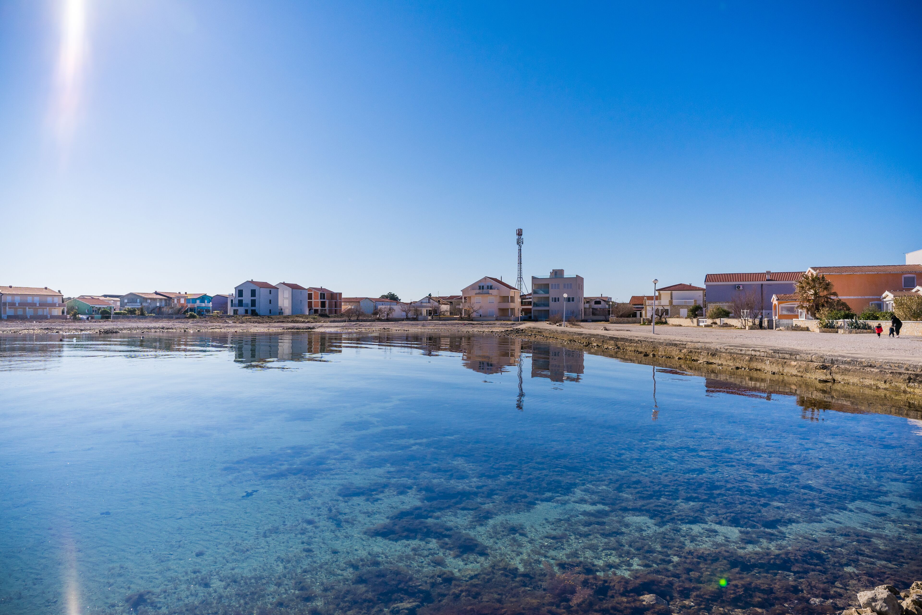 Una propiedad frente al agua con aguas cristalinas que reflejan edificios, un paseo marítimo pavimentado y algunas personas a lo lejos.