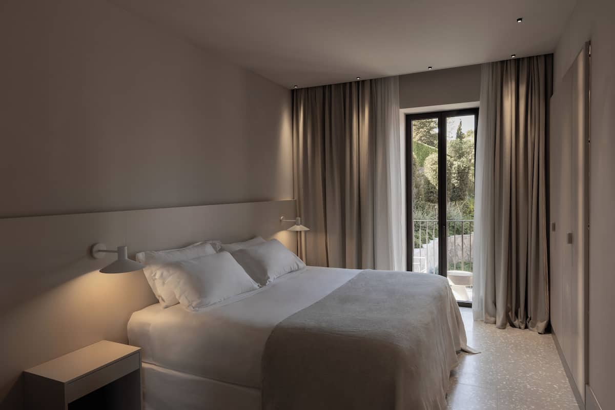 A modern hotel room with a large bed, a bedside table, and a view of the outdoors through a sliding door.