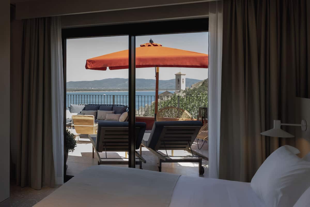 A hotel room with a balcony, a bed, two lounge chairs, an orange umbrella, and a view of the sea and mountains.