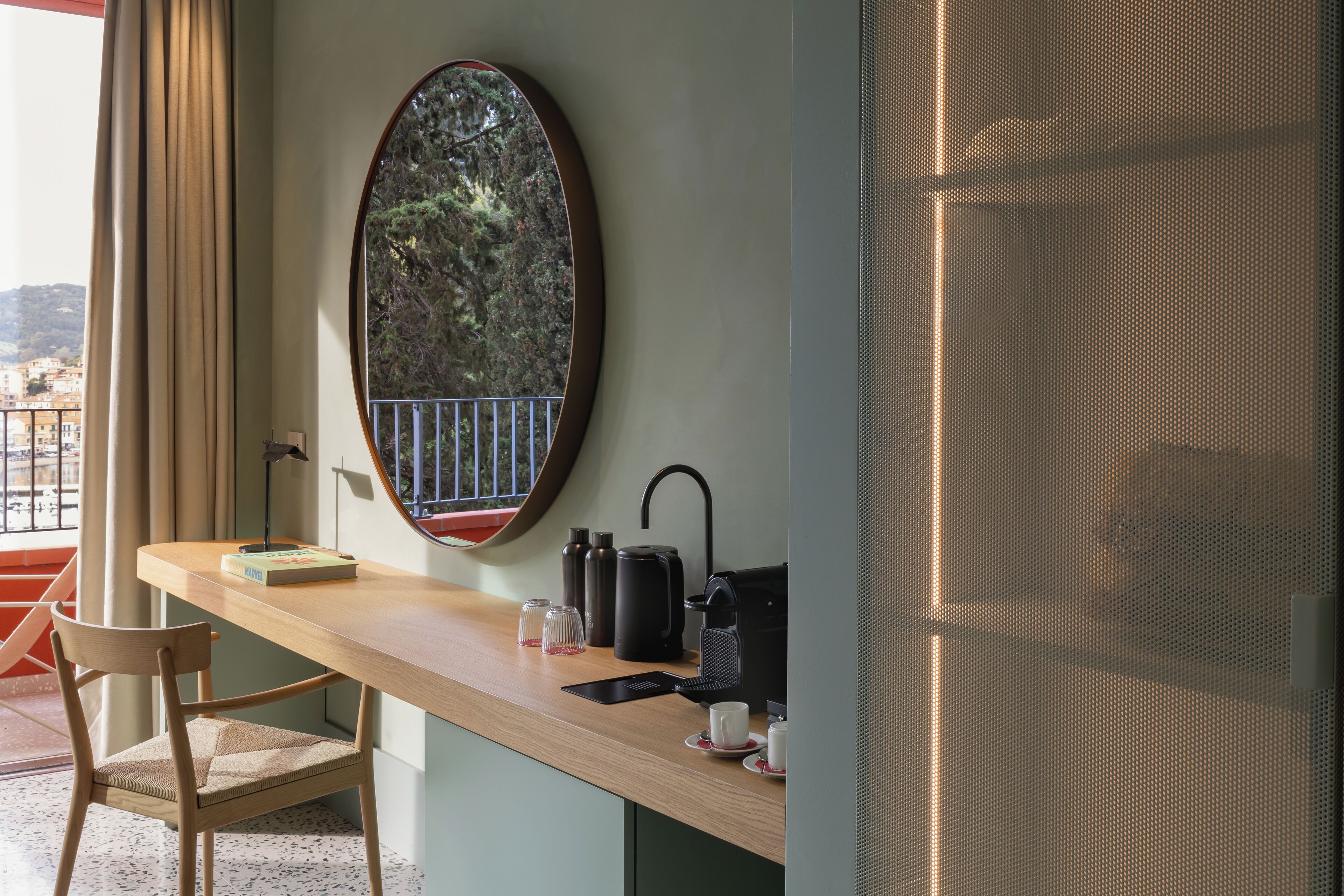 A room with a wooden desk, a chair, a round mirror, a window with a view of trees and buildings, and a door with a metal screen.