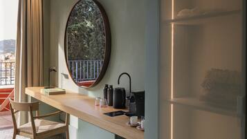 A room with a wooden desk, a chair, a round mirror, a window with a view of trees and buildings, and a door with a metal screen.