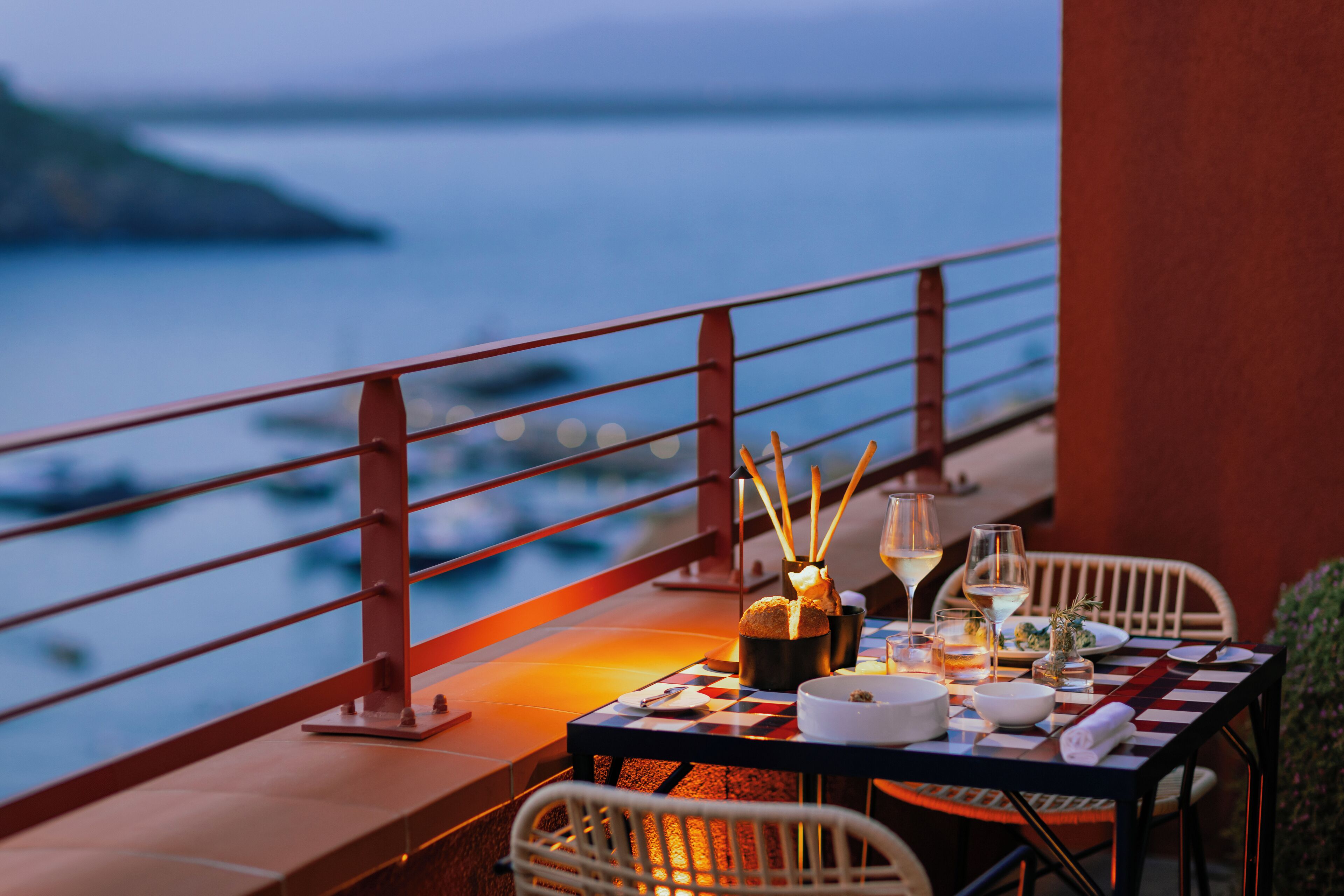A dining table set for two with a view of the sea, a red railing, and a checkered tablecloth.
