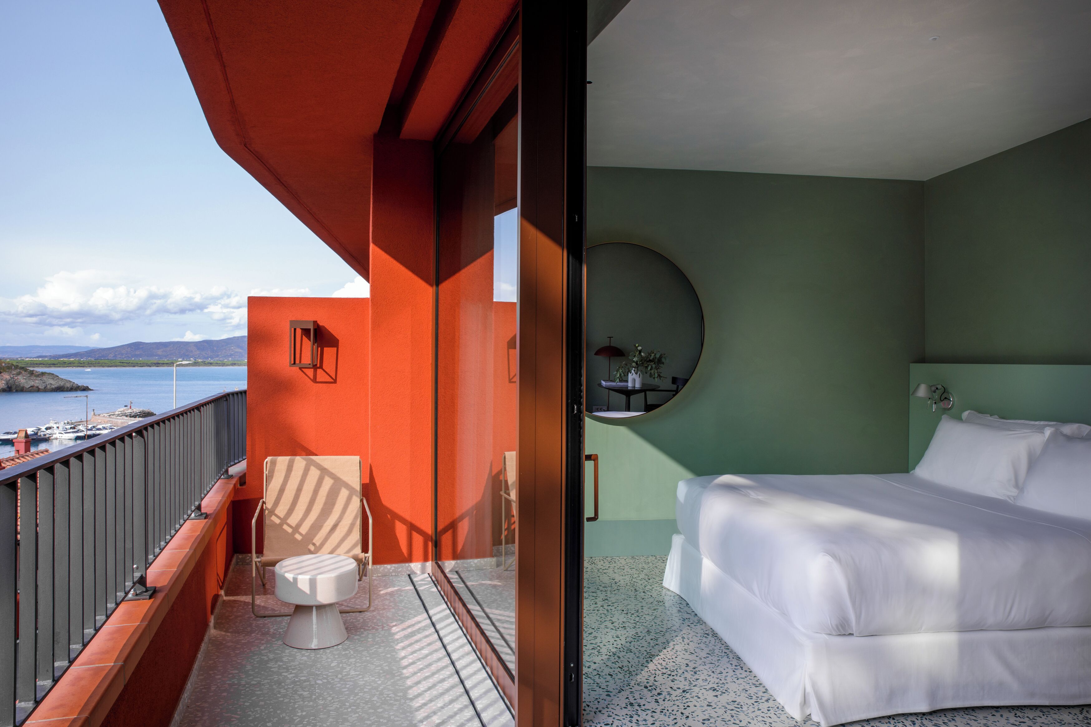 A balcony with a toilet and chair, a bed with white bedding, and a round mirror on the wall.
