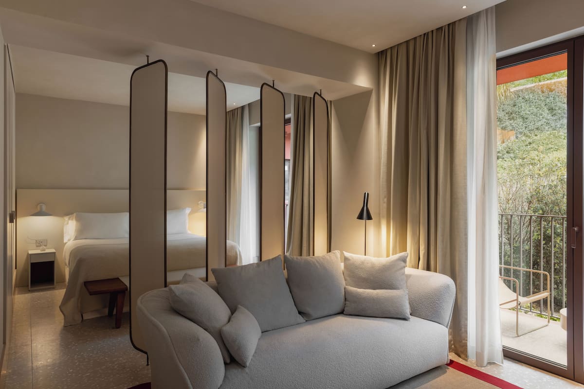 A modern hotel room with a sofa, a bed, a bedside table, and a view of the outdoors through a balcony.