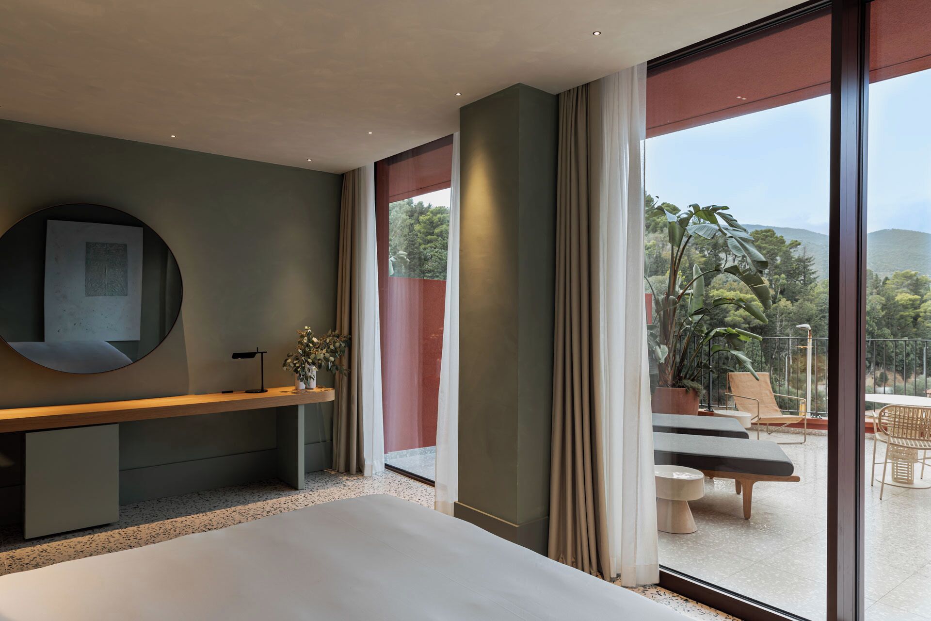 Una habitación de hotel moderna con una cama grande, una mesita de noche con lámpara, un espejo y vistas al exterior a través de amplios ventanales.