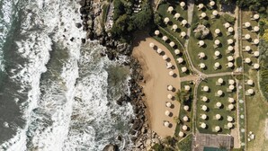 Vista aérea de un resort frente a la playa, con un litoral rocoso, una playa arenosa y una serie de mesas redondas con sombrillas dispuestas en círculo.