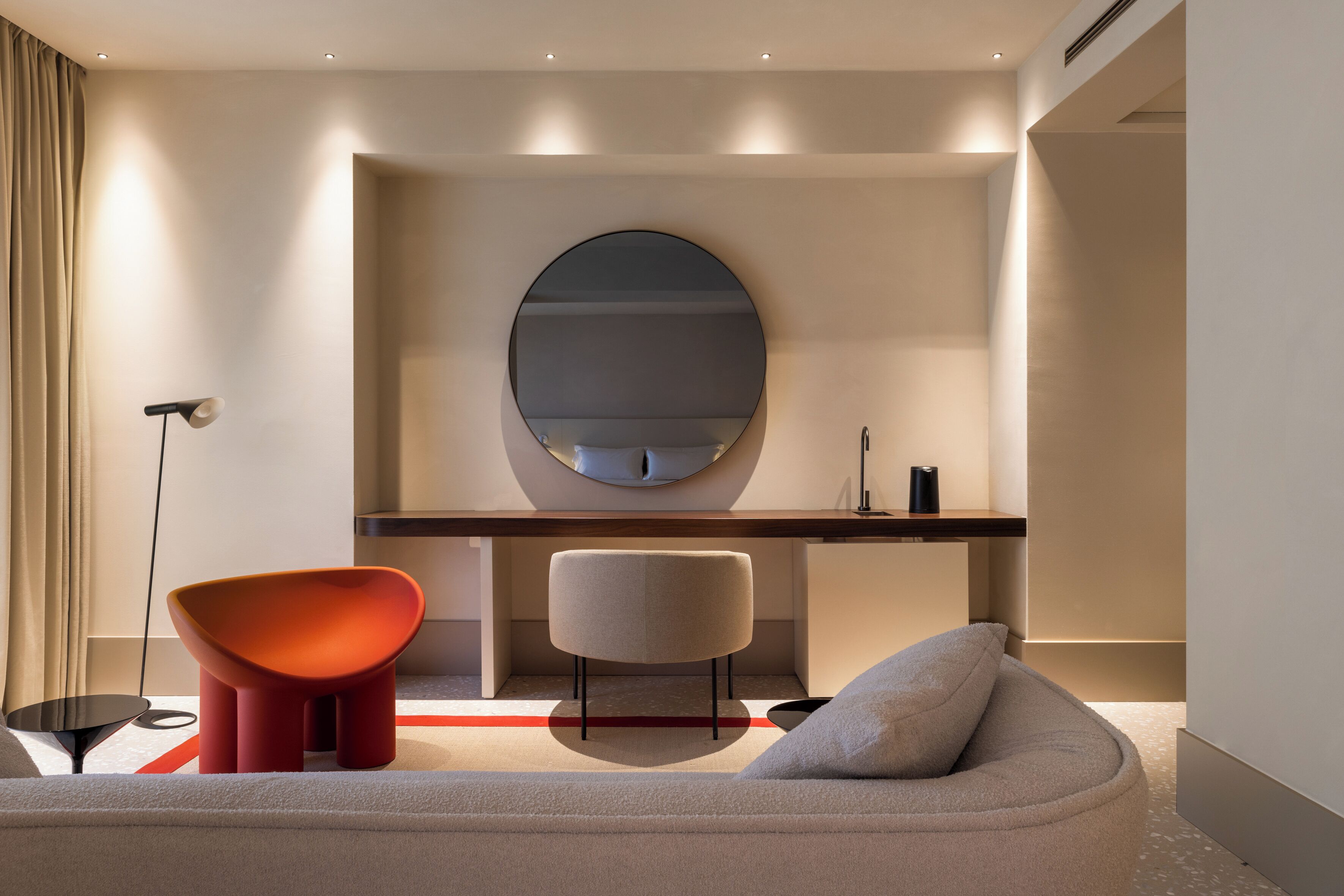 Ein modernes Hotelzimmer mit einem großen runden Spiegel, einem Sofa, einem roten Sessel und einem Schreibtisch mit einer Lampe.