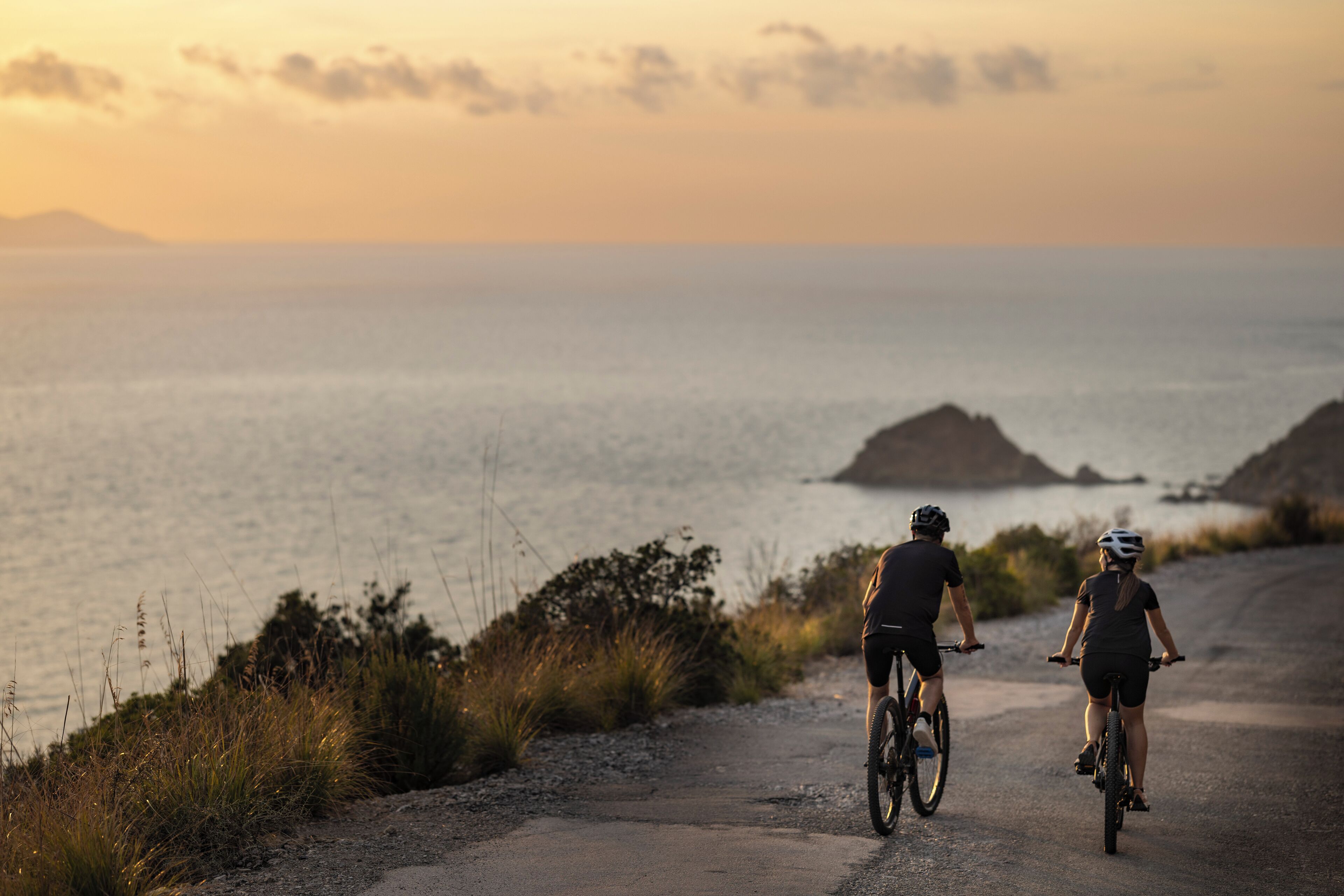 Deux cyclistes sur un chemin de gravier, avec vue sur la mer et des îles au loin.