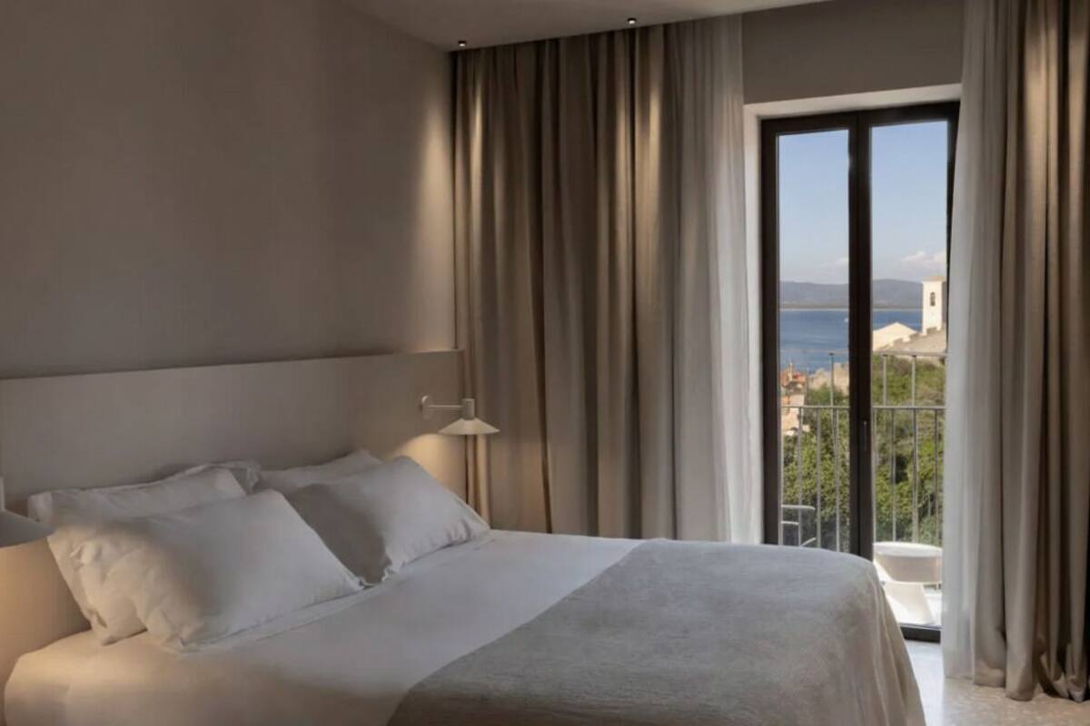 A hotel room with a large bed, a nightstand, a lamp, and a view of the sea through a sliding glass door.