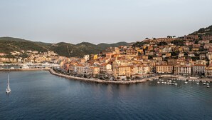 Un pueblo costero con edificios agrupados a lo largo del malecón, un puerto deportivo con barcos y colinas al fondo.