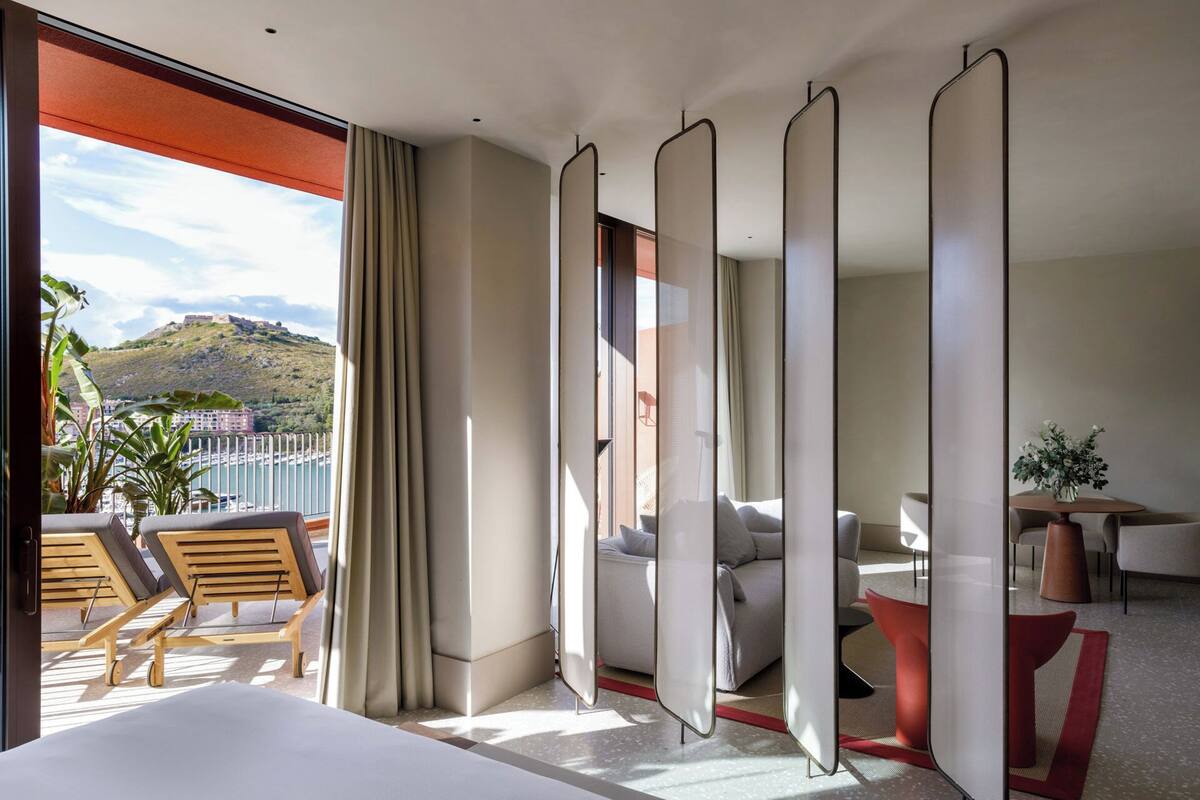 A hotel room with a balcony, a bed, a seating area with chairs, and a view of a hill.