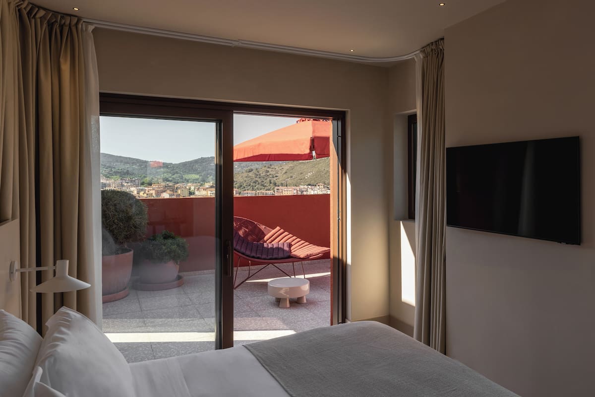 A hotel room with a bed, a TV mounted on the wall, a balcony with a red umbrella, and a view of the city.