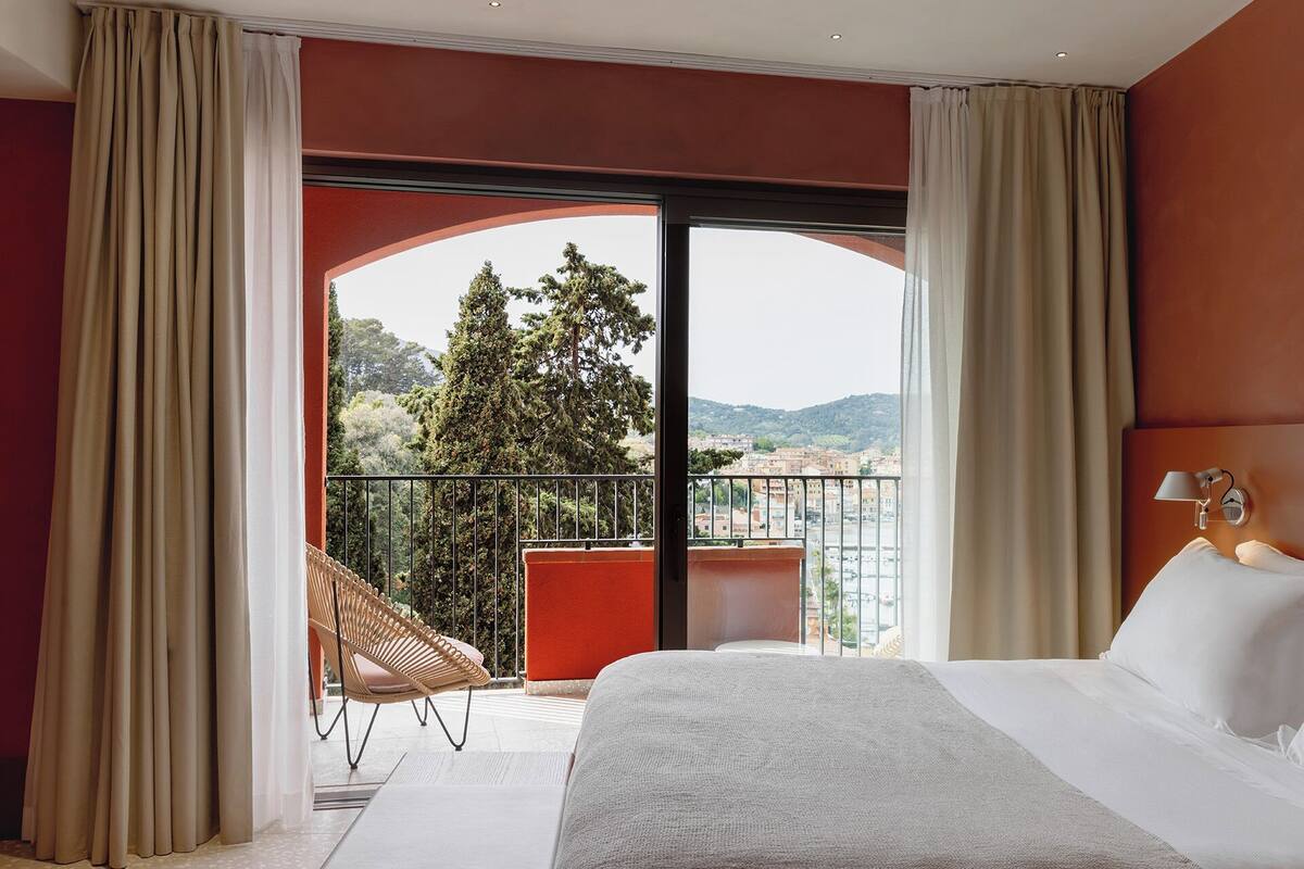 A hotel room with a large bed, a chair, and a balcony with a view of trees and buildings.