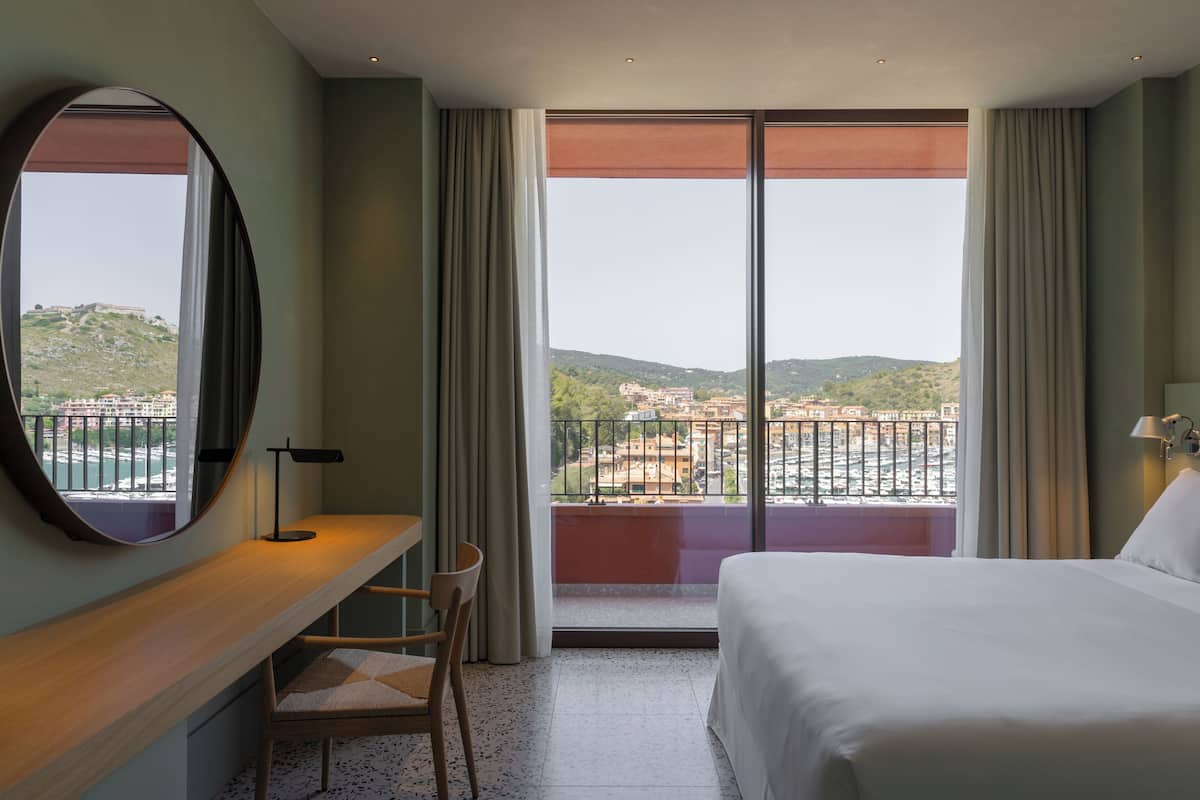 A hotel room with a bed, a desk with a chair, a large window with curtains, and a view of the city.