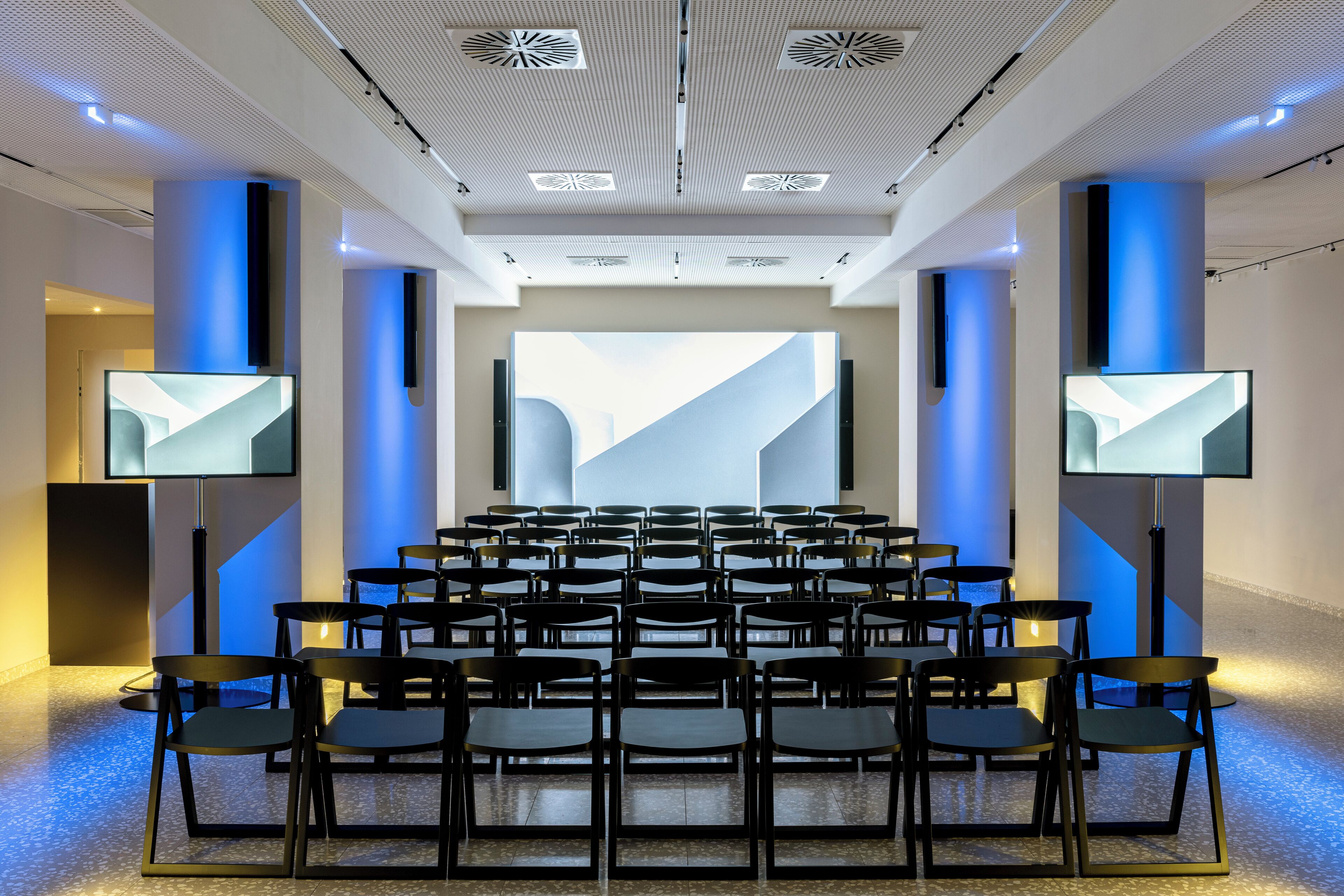 Ein Konferenzraum mit Reihen von Stühlen, einem großen Bildschirm an der Vorderseite und modernem Deckenstil.