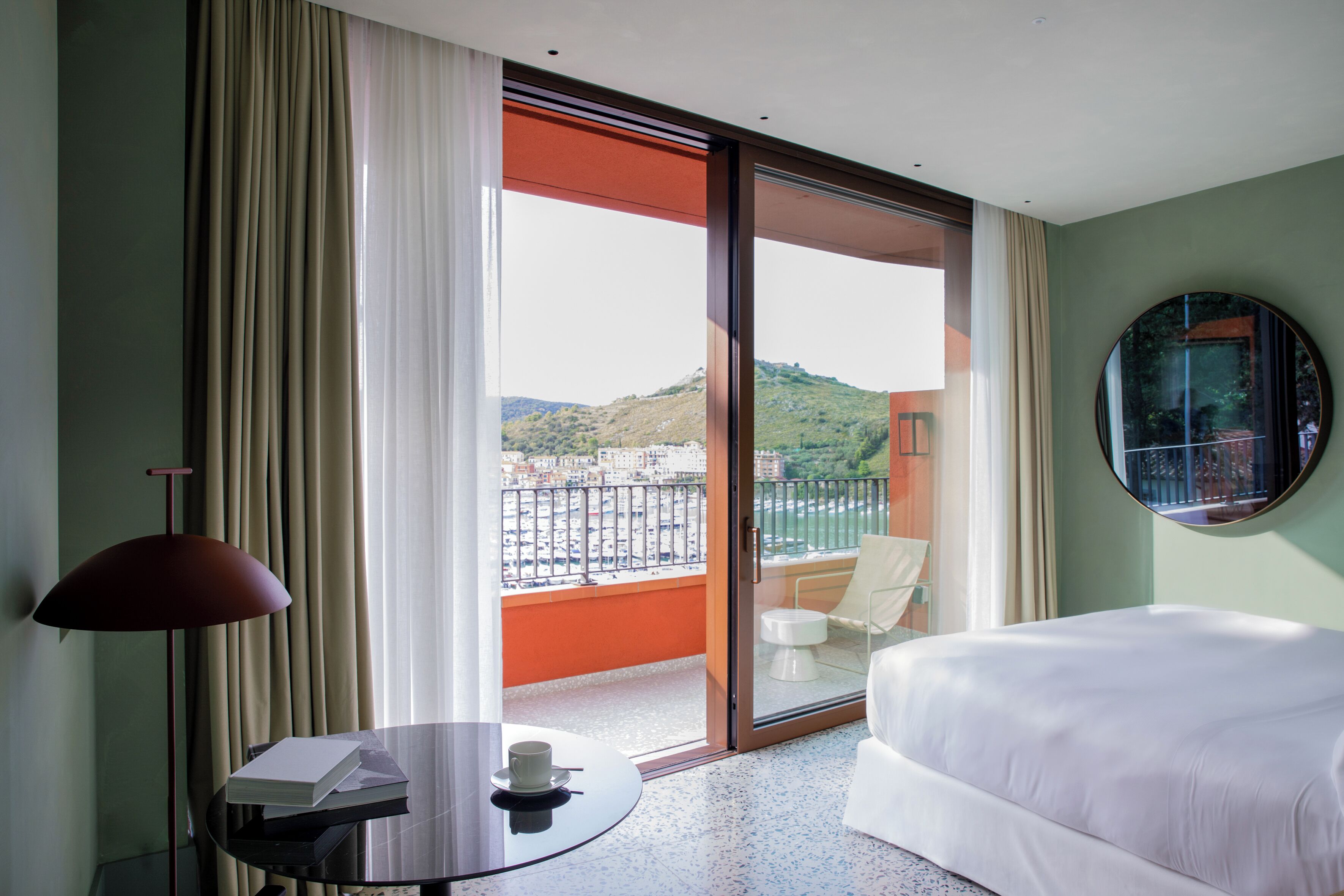 A hotel room with a bed, a table with a cup and book, a chair, and a view of a balcony with a railing and a chair.