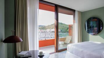 A hotel room with a bed, a table with a cup and book, a chair, and a view of a balcony with a railing and a chair.