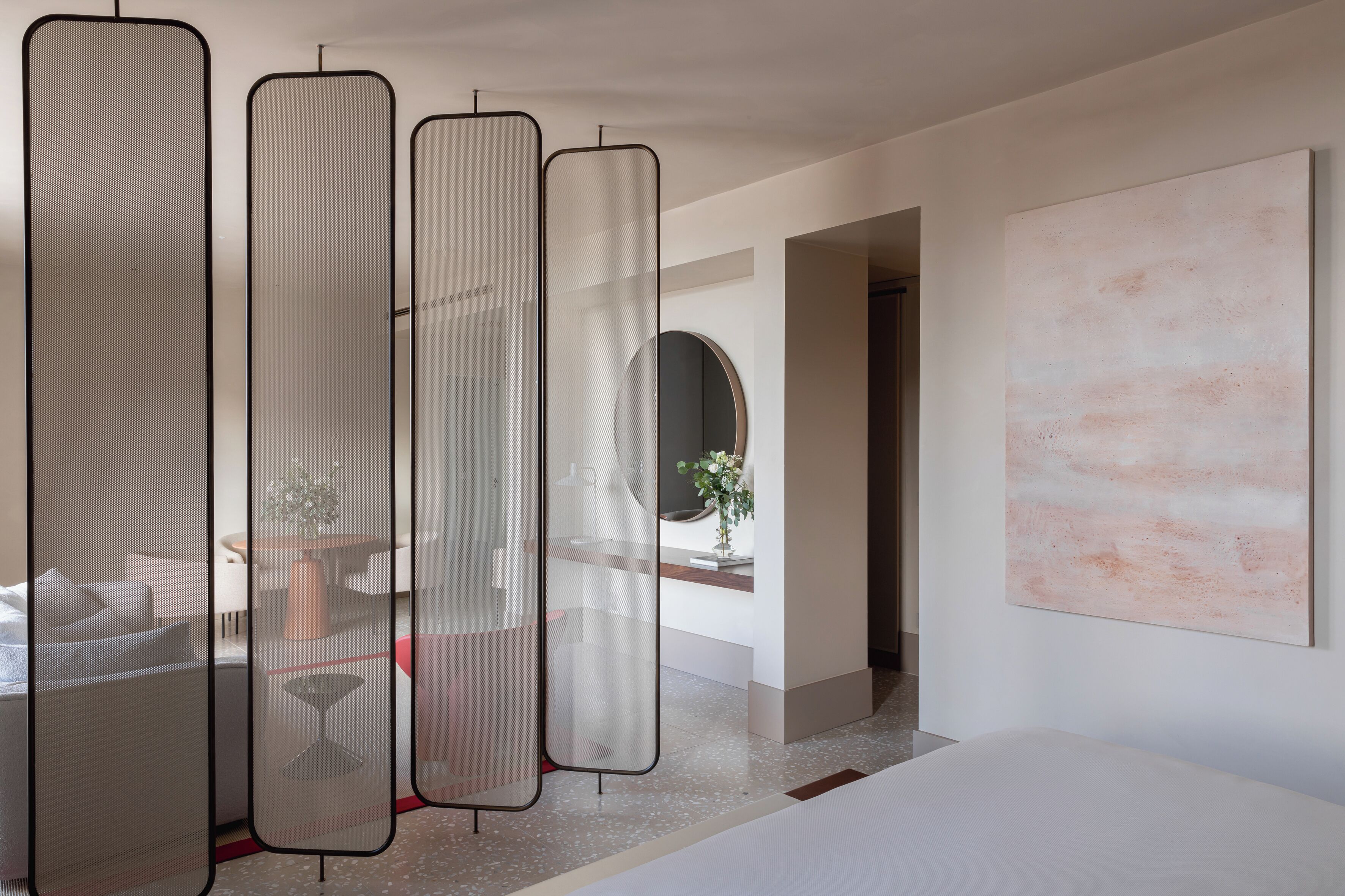 Een moderne hotelkamer met een groot bed, een ronde spiegel, een rond tafeltje met een vaas en een groot abstract schilderij aan de muur.