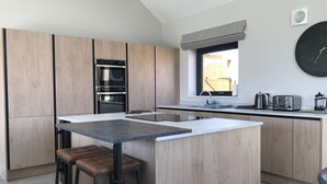 Een moderne keuken met houten kastjes, een inbouwoven, een groot kookeiland met barkrukken, een raam met uitzicht en een ronde klok aan de muur.