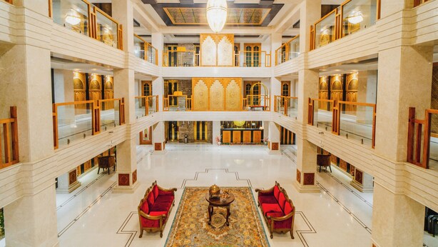 A spacious lobby with a high ceiling, ornate chandeliers, and a central seating area with red upholstered chairs. - Hotel The Dewan (Srinagar)