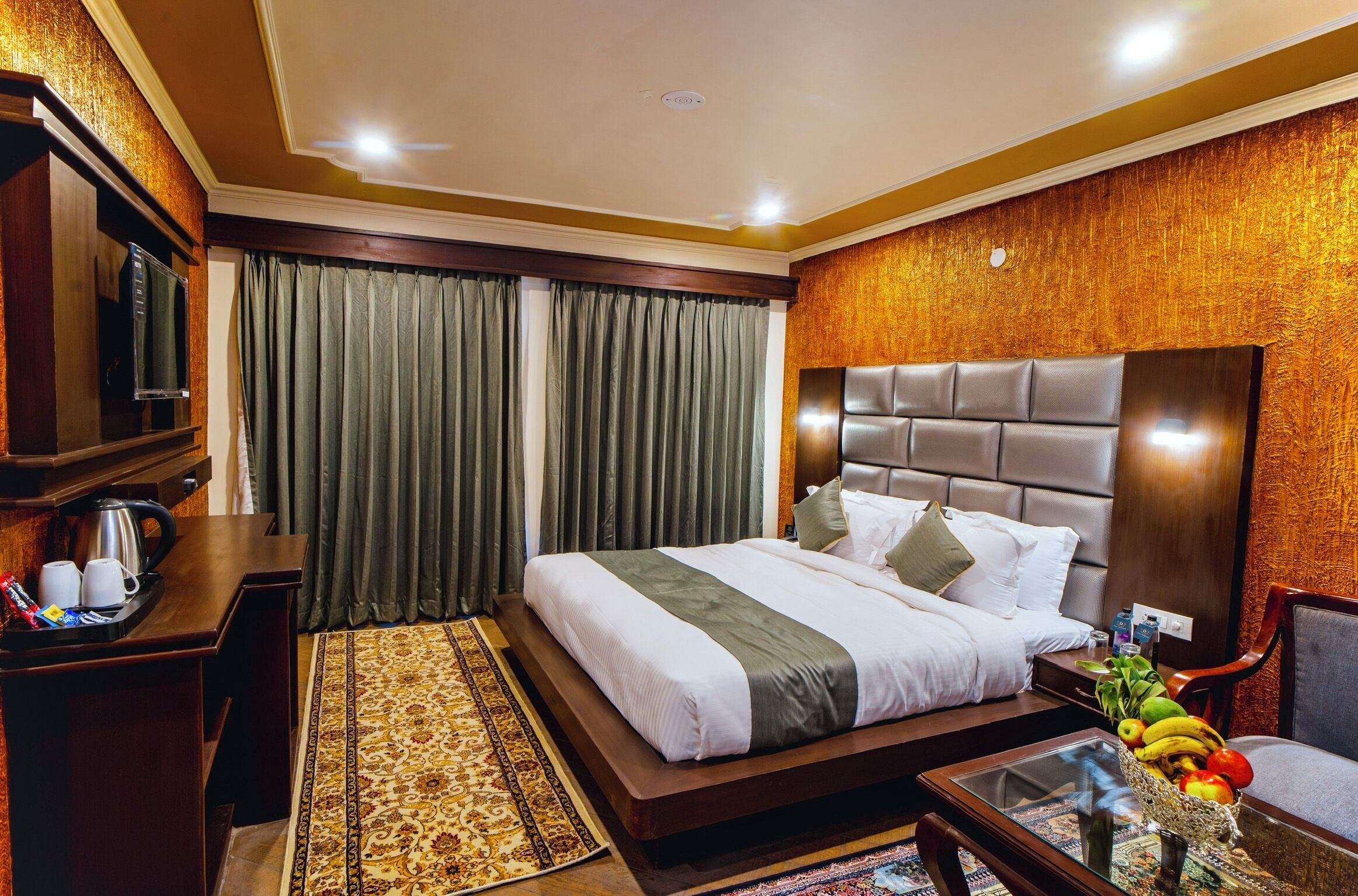 Quarto de hotel com uma cama grande, cabeceira de madeira, escrivaninha com chaleira, bandeja de café e cesta de frutas na mesa lateral.