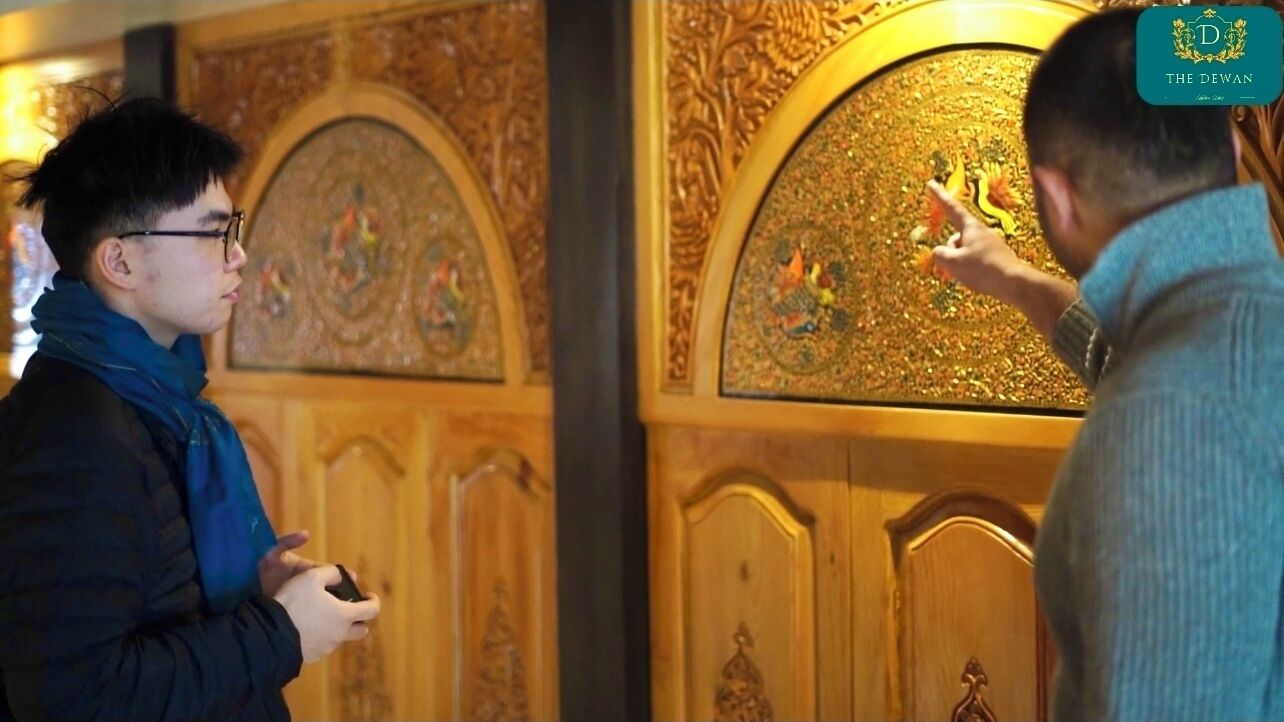 Two individuals in a room with ornate wooden doors and detailed wall panels.
