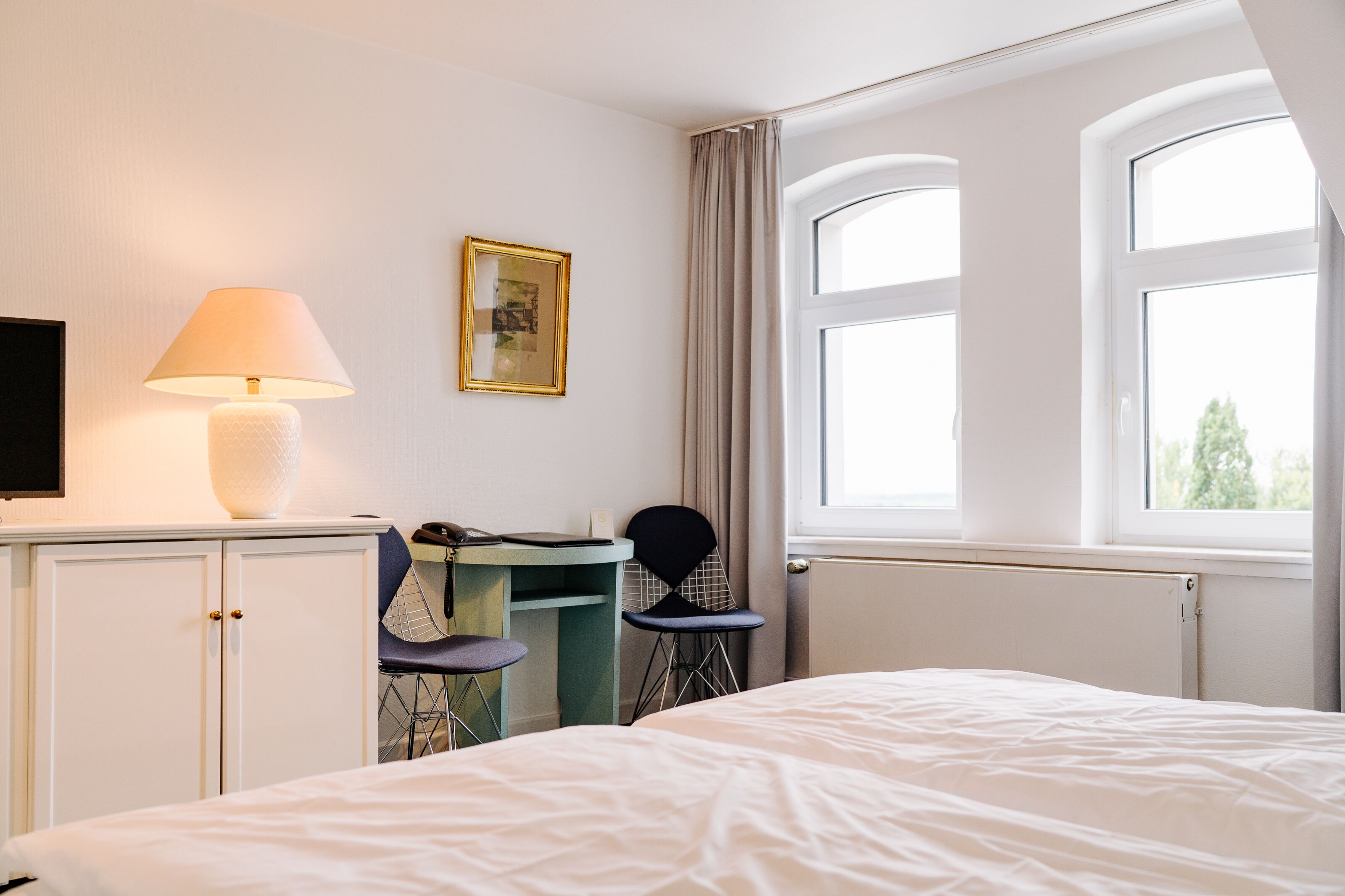 A hotel room with a bed, a desk with a chair, a lamp, a television, and a window with curtains.