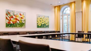 A conference room with a long table and chairs, two paintings on the walls, and a large window with curtains.