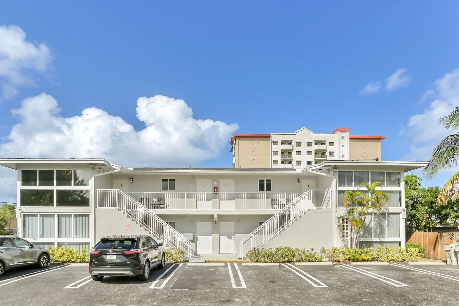 A two-story hotel with a central staircase, parking lot, and a building with a red roof.