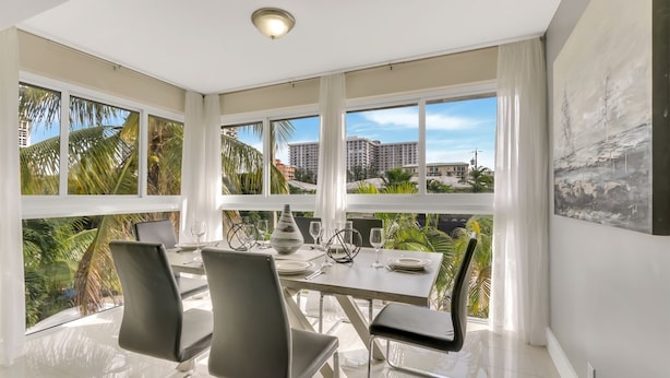 A modern dining area with a large table, four chairs, and a view of palm trees and buildings through large windows. - RiversideStar: Pompano Retreat, close to beach (Pompano Beach)