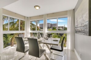 A modern dining area with a large table, four chairs, and a view of palm trees and buildings through large windows. - RiversideStar: Pompano Retreat, close to beach (Pompano Beach)