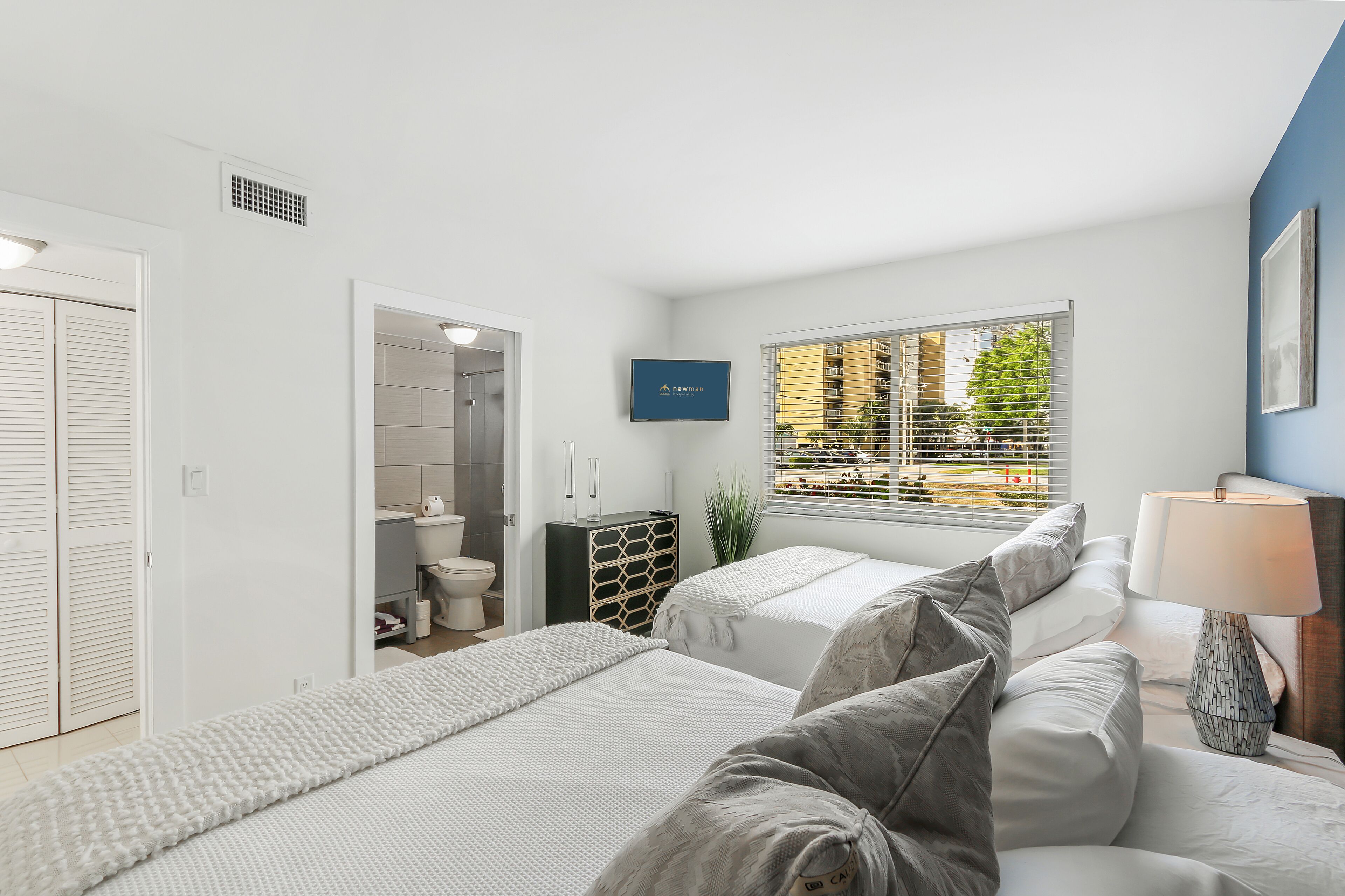 A modern bedroom with two beds, a TV, a window with blinds, and a view of the outside.