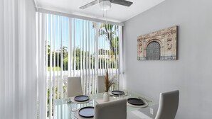 A modern dining area with a glass table, white chairs, and a view of greenery outside.