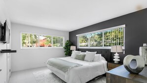 A modern bedroom with a large bed, a nightstand, a TV, and a view of greenery outside.