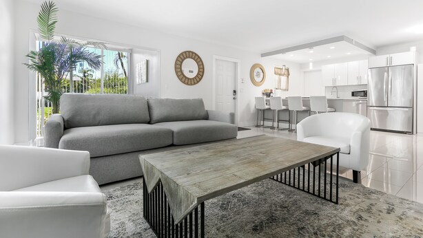 A modern living room with a grey sofa, white armchairs, a wooden coffee table, and a kitchen in the background.