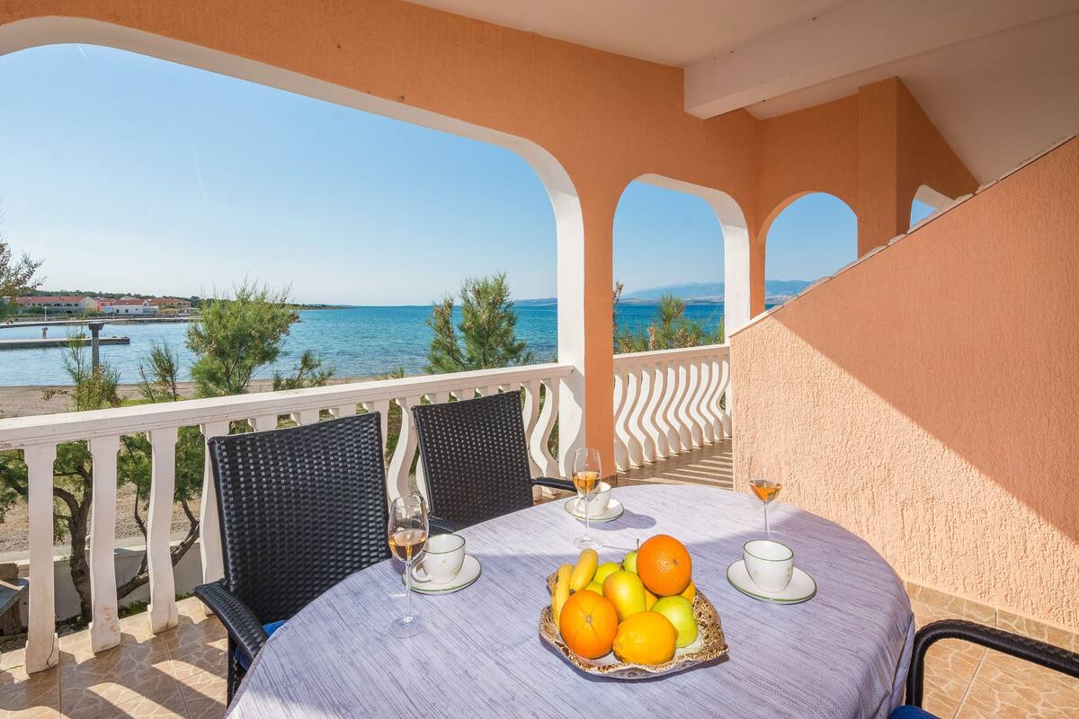 Un balcón con una mesa dispuesta para dos personas, con una cesta de frutas, dos copas de vino y vistas al mar y edificios.