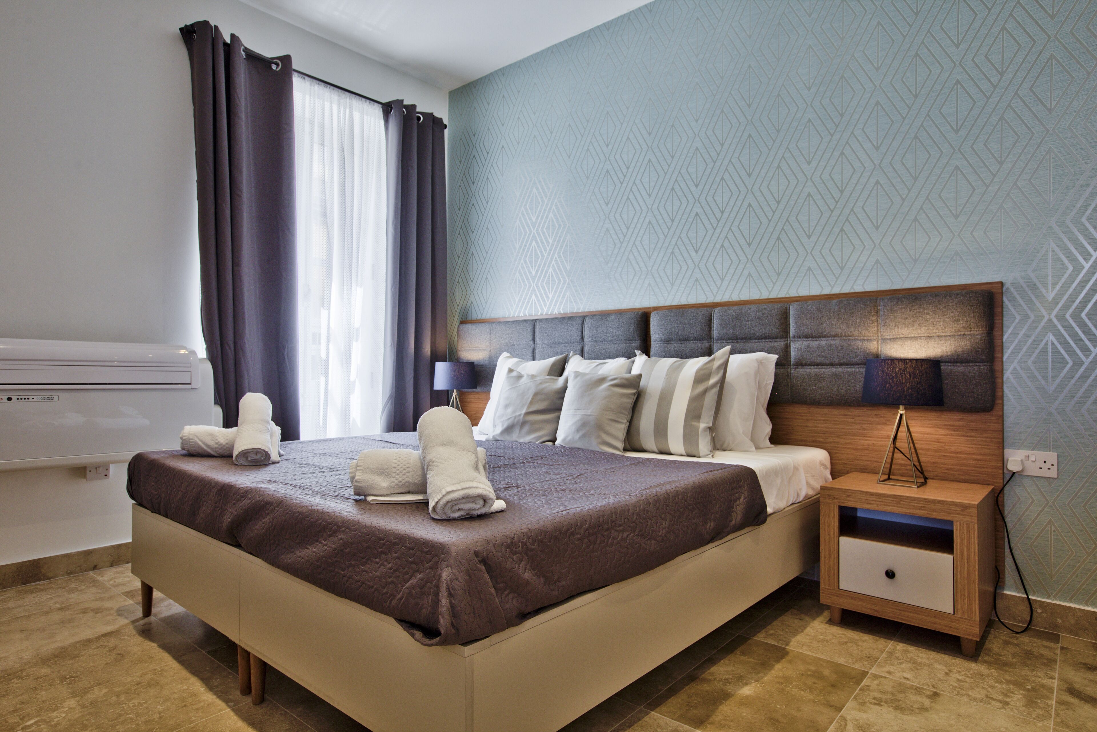 Una cama bien hecha con cubrecolcha marrón, una mesita de noche con lámpara y una pared con diseño geométrico.