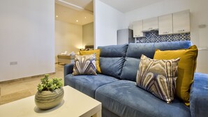 A modern living room with a blue sofa, a small table with a plant, and a kitchen in the background. - Ursula Suites- Self Catering Apartments- Valletta- by Tritoni Hotels (Valletta)
